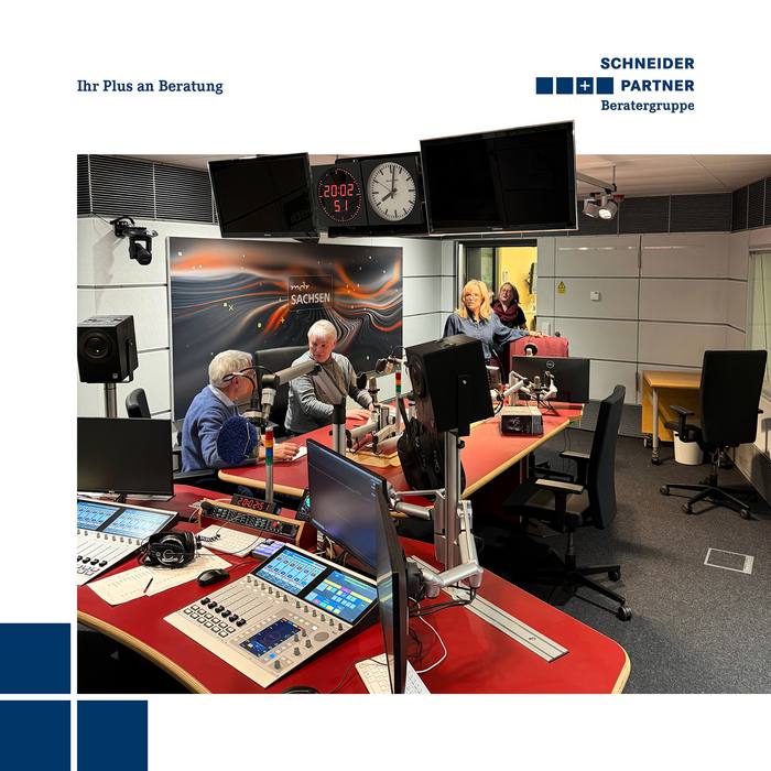 Blick in ein Aufnahmestudio mit verschiedener Tontechnik und Bildschirmen im Vorder- und einigen Personen im Hintergrund.